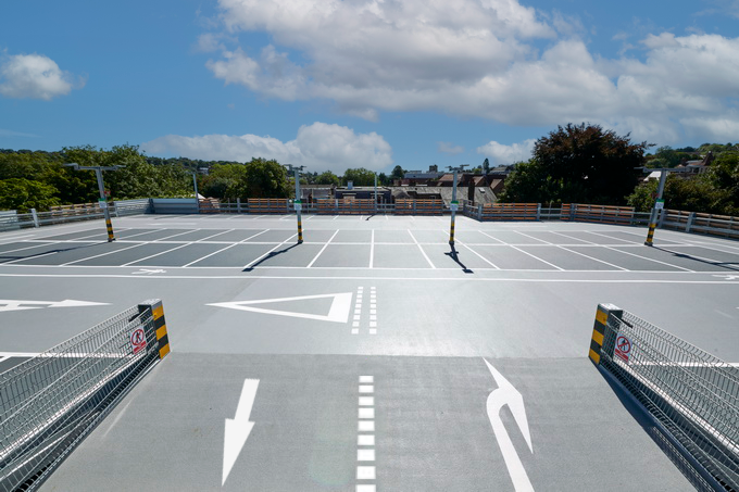Ein leerer überdachter Parkplatz mit klaren weißen Markierungen und Pfeilen, umgeben von einem Zaun, unter einem teilweise bewölkten Himmel mit Bäumen und Gebäuden im Hintergrund.